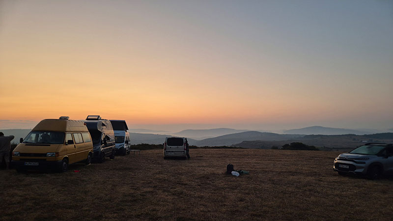 Mehrere Camper und Autos stehen auf einer trockenen Wiese in der hügeligen Landschaft Nordspaniens. Am Horizont leuchtet der Himmel orange, während sich in den Tälern bereits Dunst oder Nebel ausbreitet.