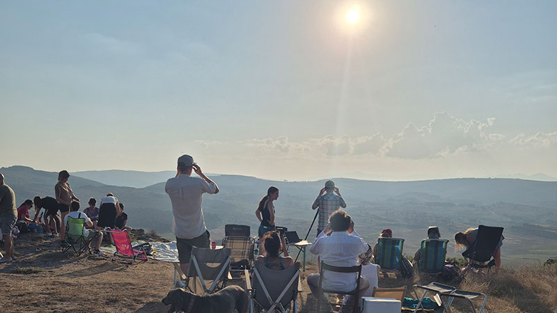 Eine Gruppe von Menschen sitzt und steht mit Campingstühlen auf einer trockenen Anhöhe und blickt zur Sonne über der weiten Hügellandschaft. Einige beobachten den Himmel mit Schutzbrillen oder Kameras, während zwei Hunde zwischen den Stühlen liegen.